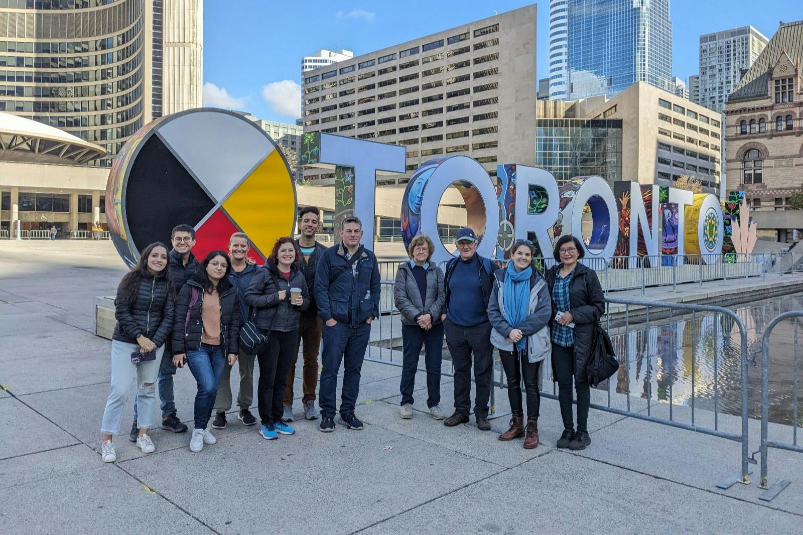 在一个拥有现代建筑的城市广场上，一群人站在一个大牌子前，牌子上写着五颜六色的字母。