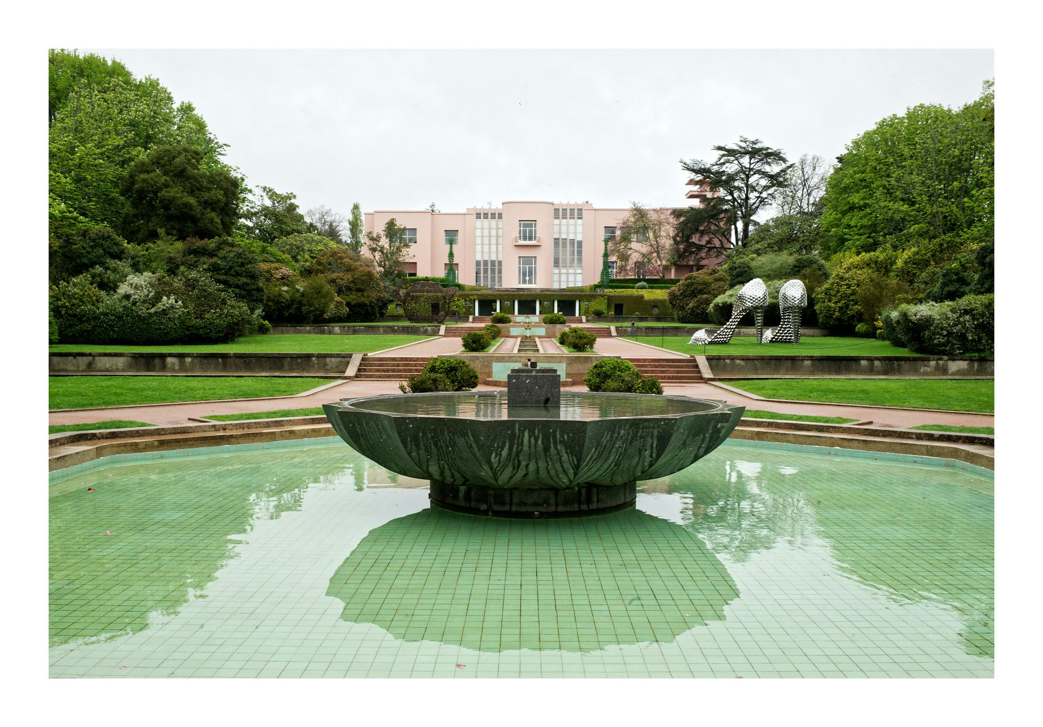 En trädgård med en fontän, en stig som leder till en rosa byggnad och stora högklackade skoskulpturer mitt i grönska.