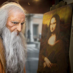 Äldre man med långt vitt hår och skägg i brun mantel står bredvid en målning av Mona Lisa.