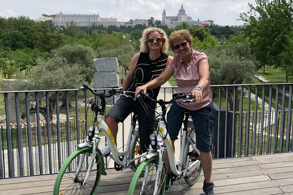 Two women with bikes smile on a bridge with a scenic park and historic buildings in the background.
