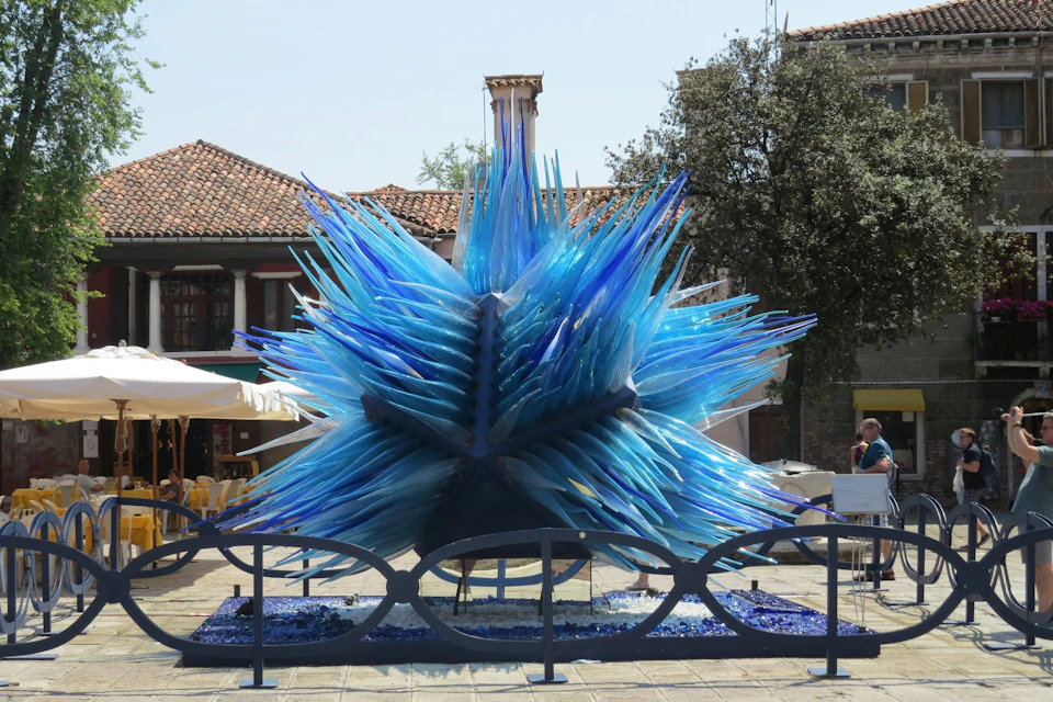 A large blue glass sculpture resembling a spiky starburst, displayed outdoors with a building and trees in the background.