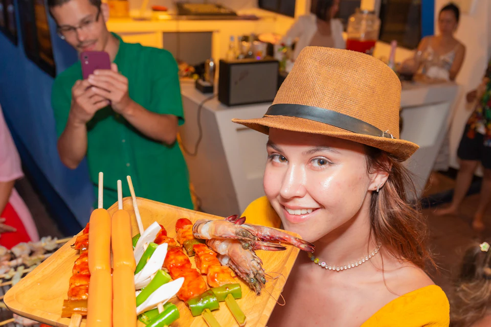 Woman in a hat holding a tray of skewers with meat, vegetables, and sausages at a lively indoor barbecue party.