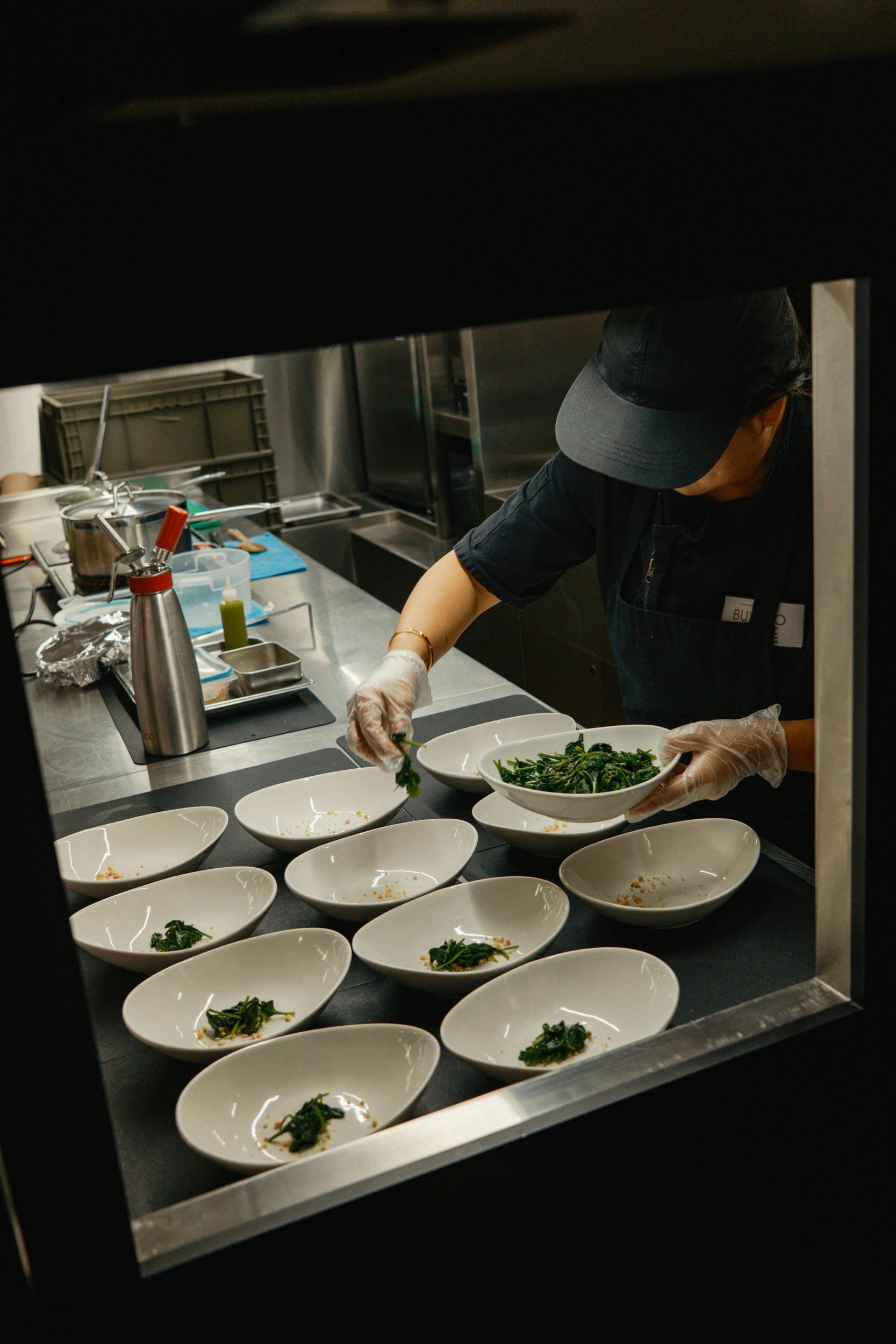 Un chef en uniforme sombre prépare plusieurs assiettes de légumes verts dans une cuisine professionnelle.