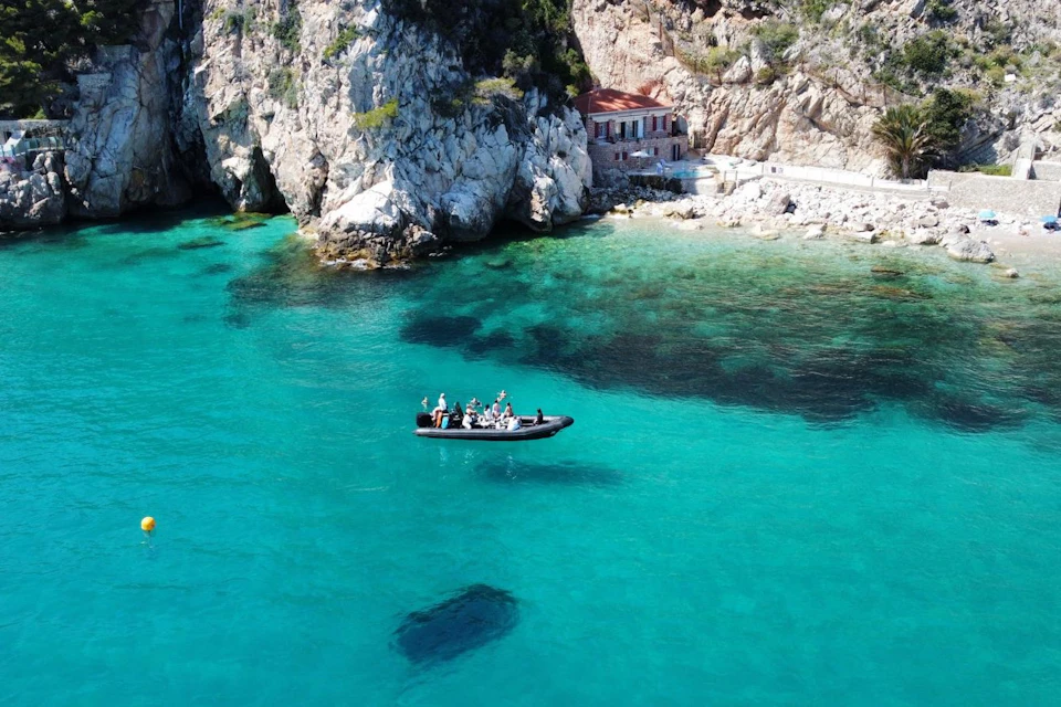 A group of people in a small motorized boat on clear turquoise water near a rocky cliff and a small building on the shore.