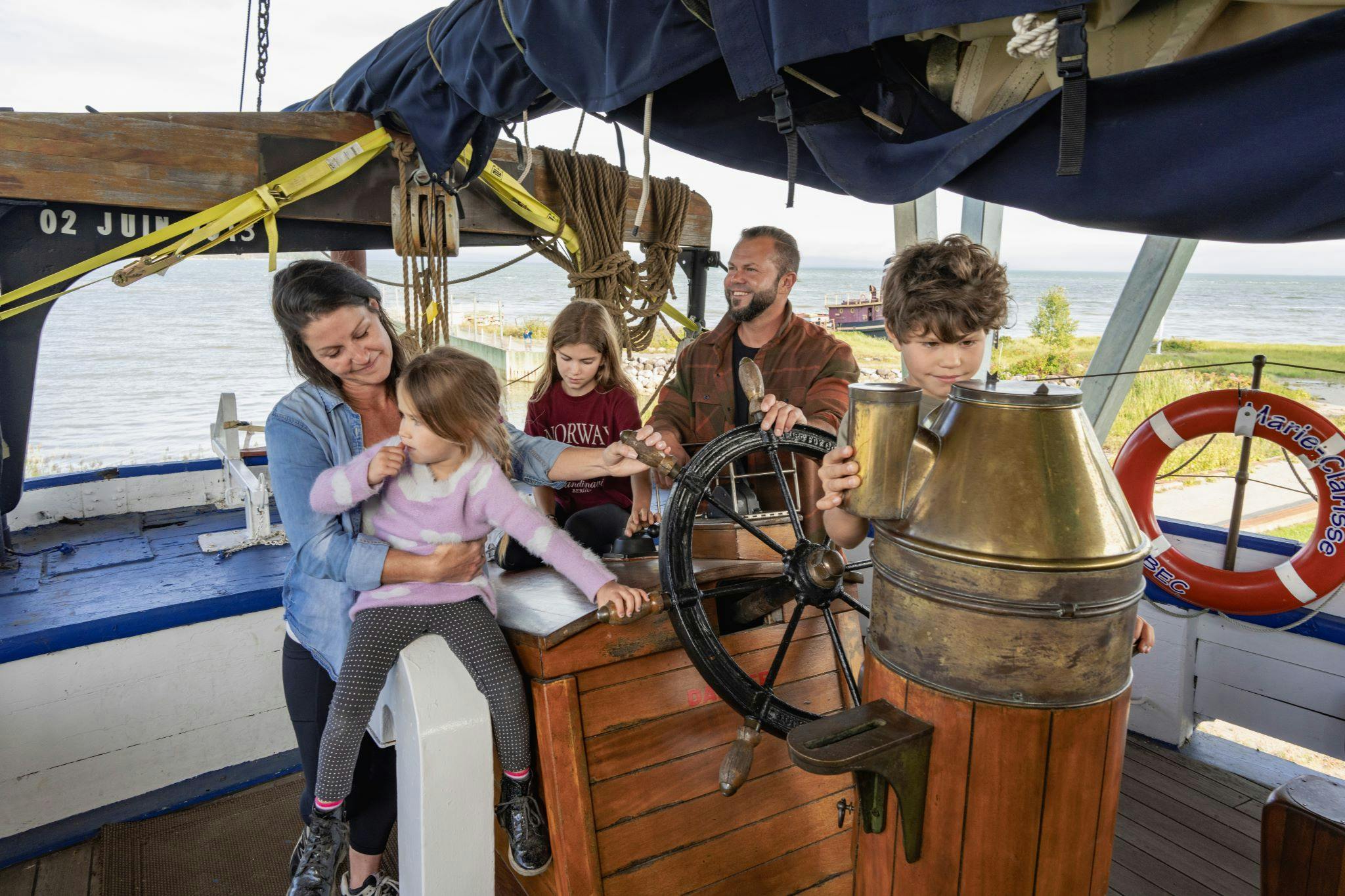 En mand og tre børn sidder ved roret på en båd, med et barns styring. På en redningsring står der "Atlanterhavskrydstogt". Havet er synligt i baggrunden.
