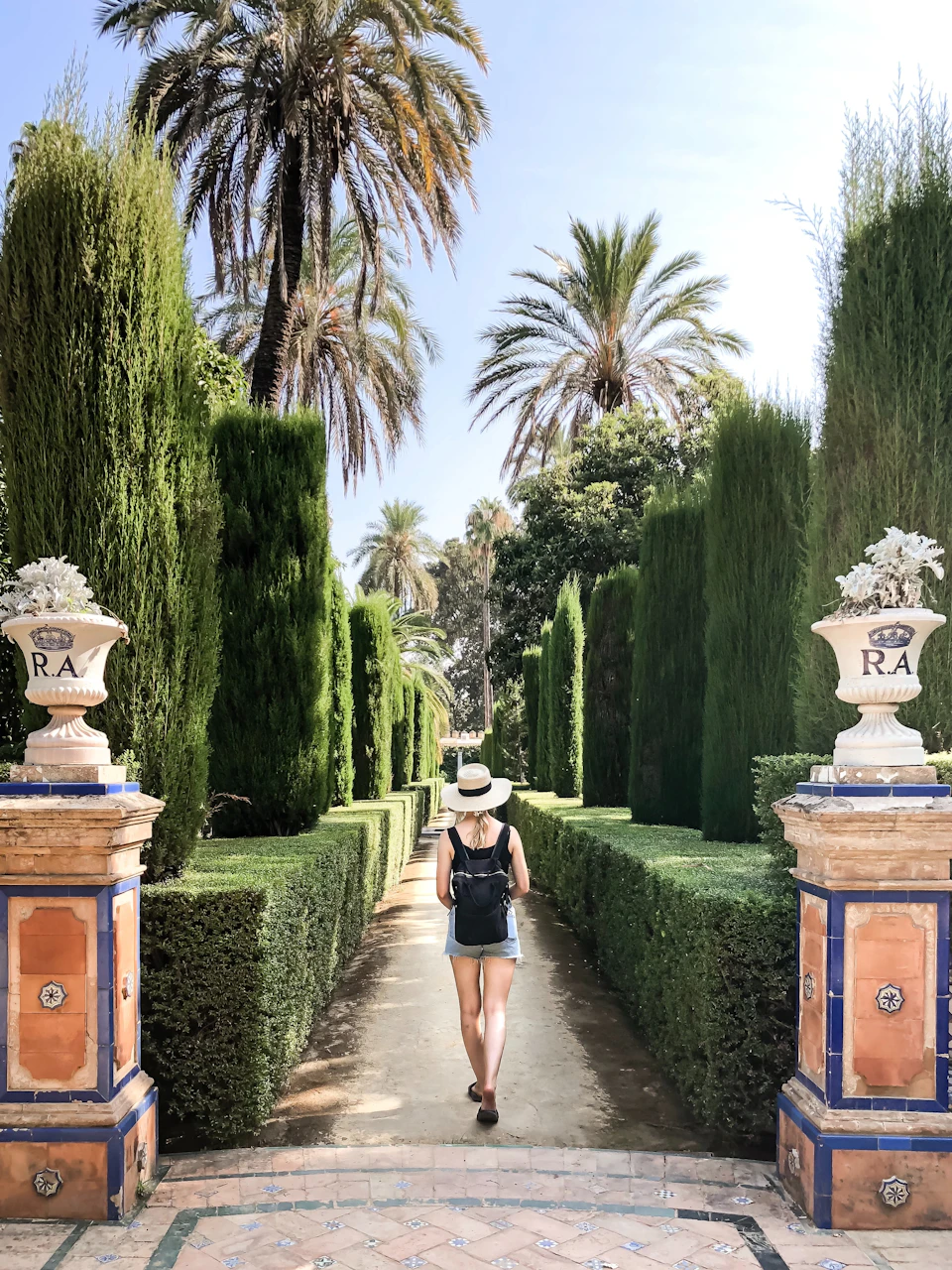 A person with a backpack and hat walks through a manicured garden path flanked by tall hedges and palm trees under a clear sky.