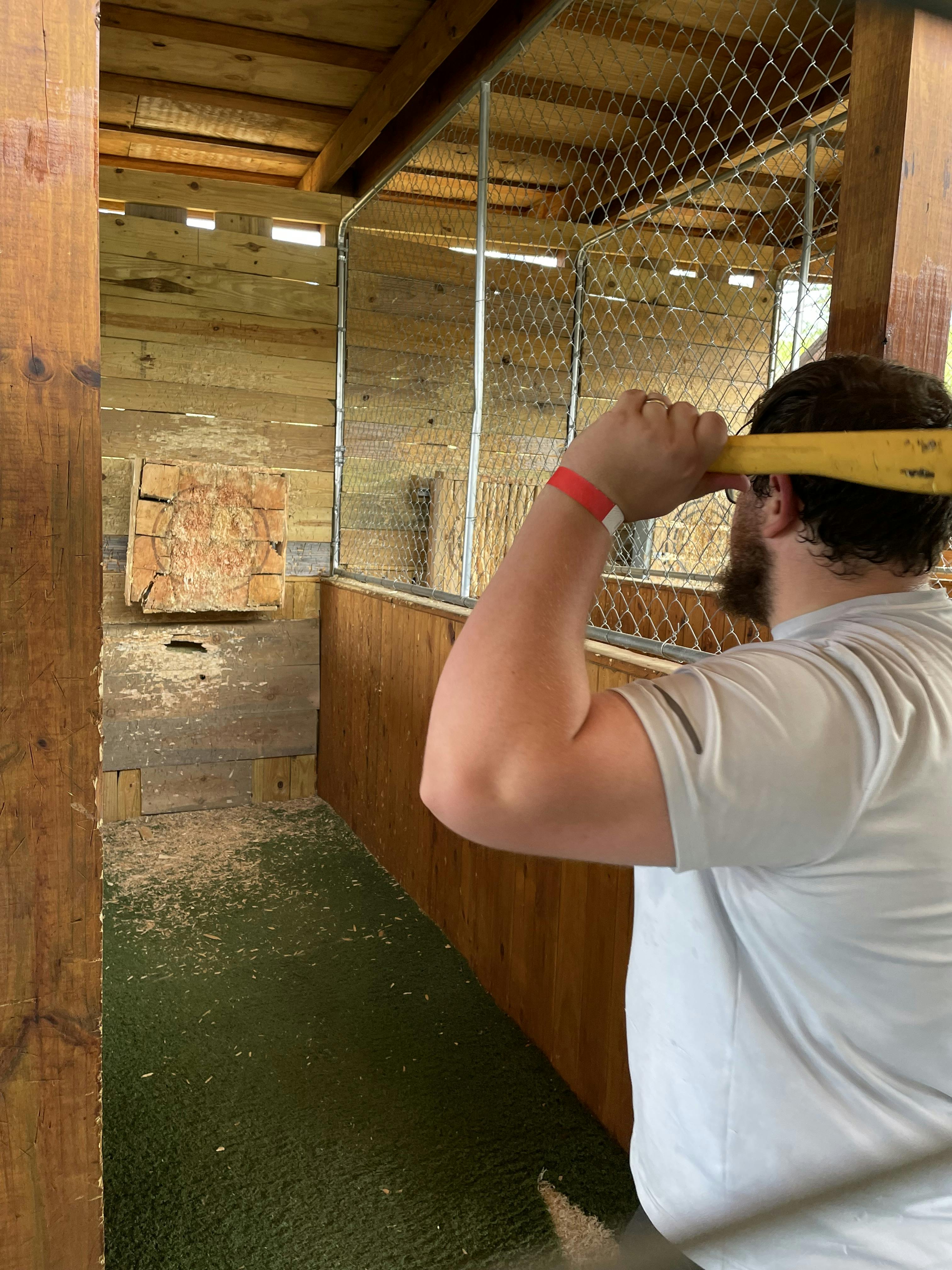 A person wearing a white shirt and a red wristband is preparing to throw an axe at a wooden target in an enclosed axe-throwing lane.