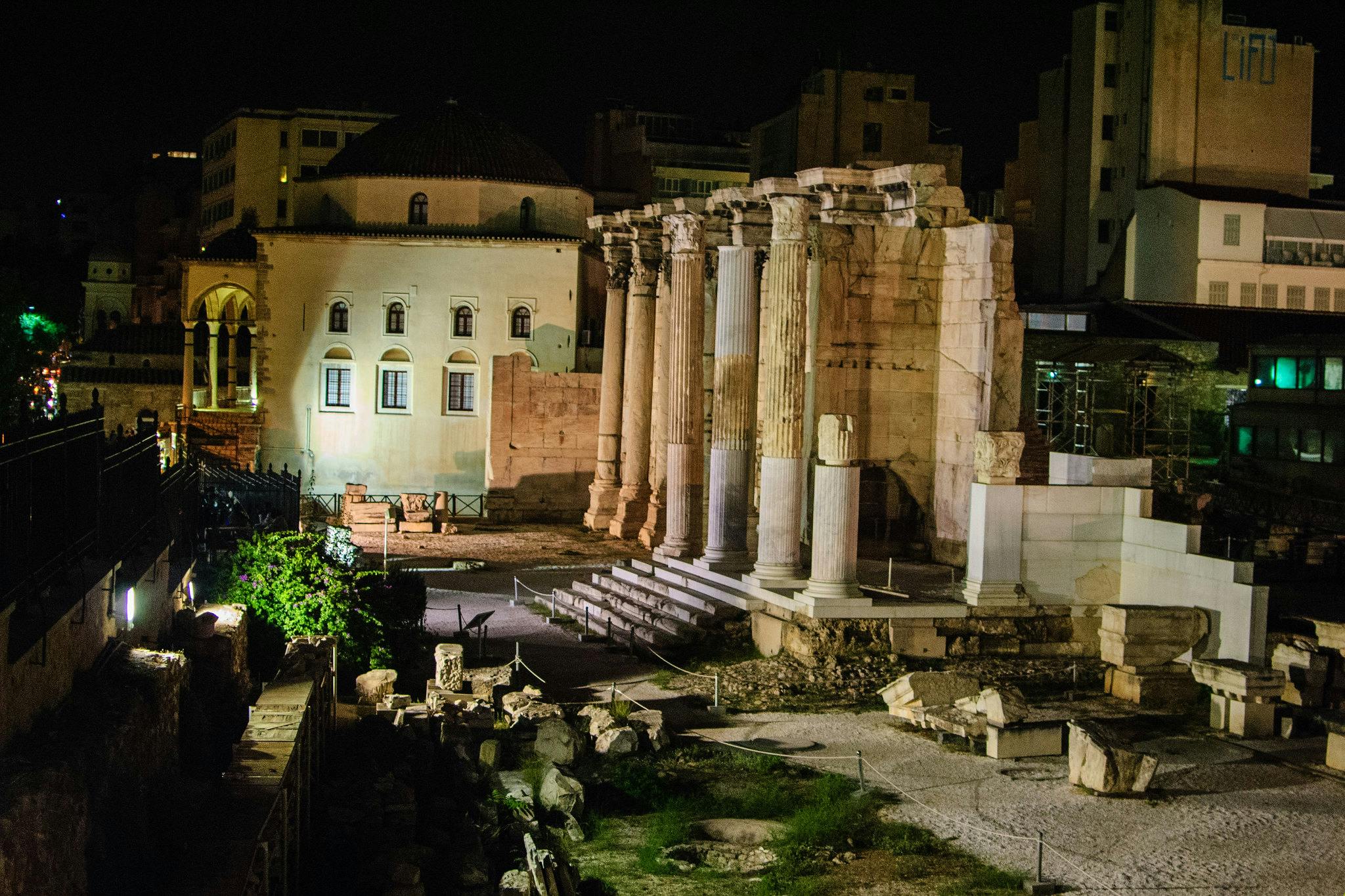 Forntida grekiska ruiner med höga pelare och en välbevarad byggnad, upplyst på natten, omgiven av moderna strukturer.