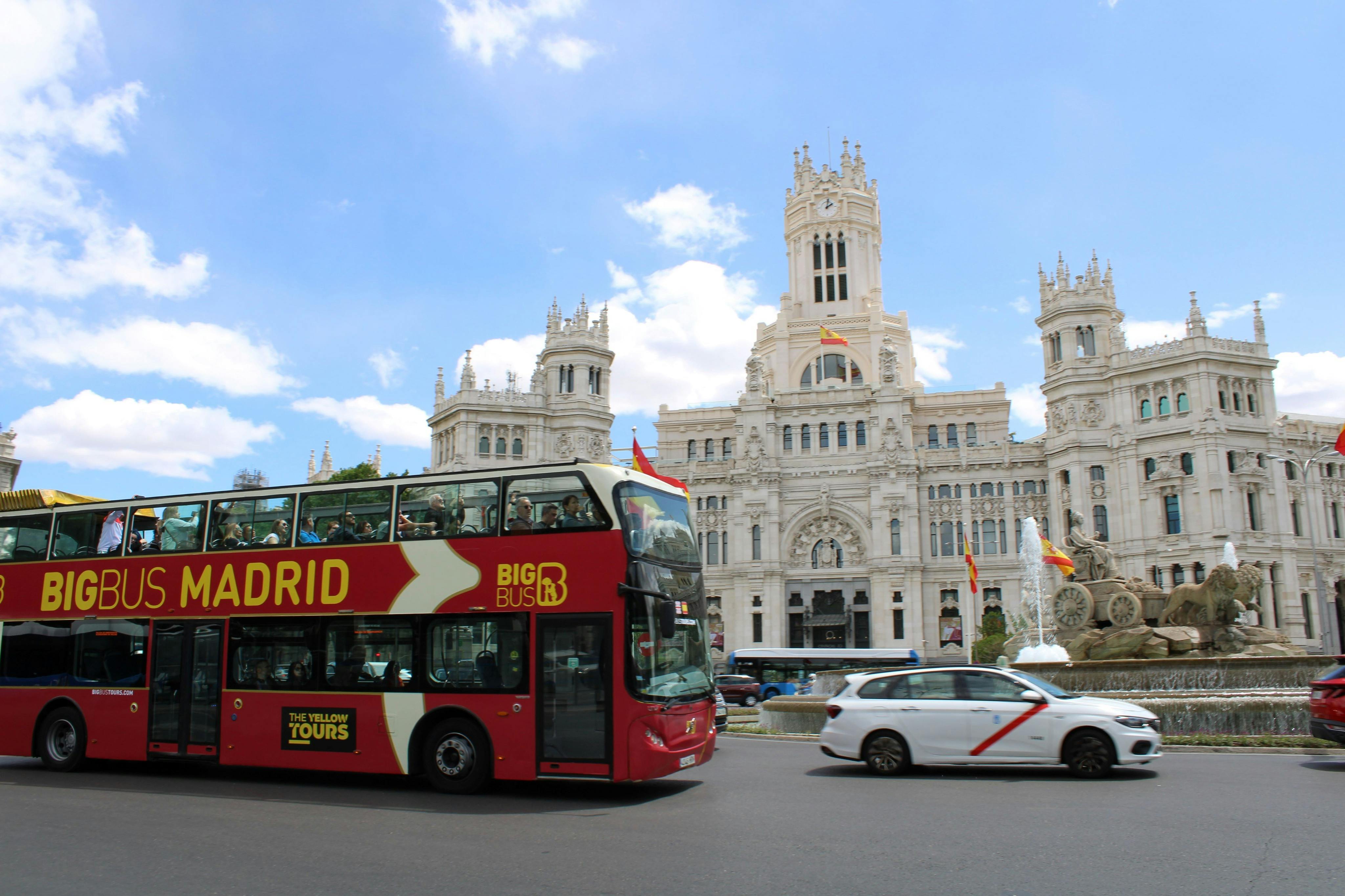 Před velkou bílou budovou s ozdobnou architekturou pod modrou oblohou projíždí červený dvoupatrový autobus s „Velkým autobusem Madrid“.