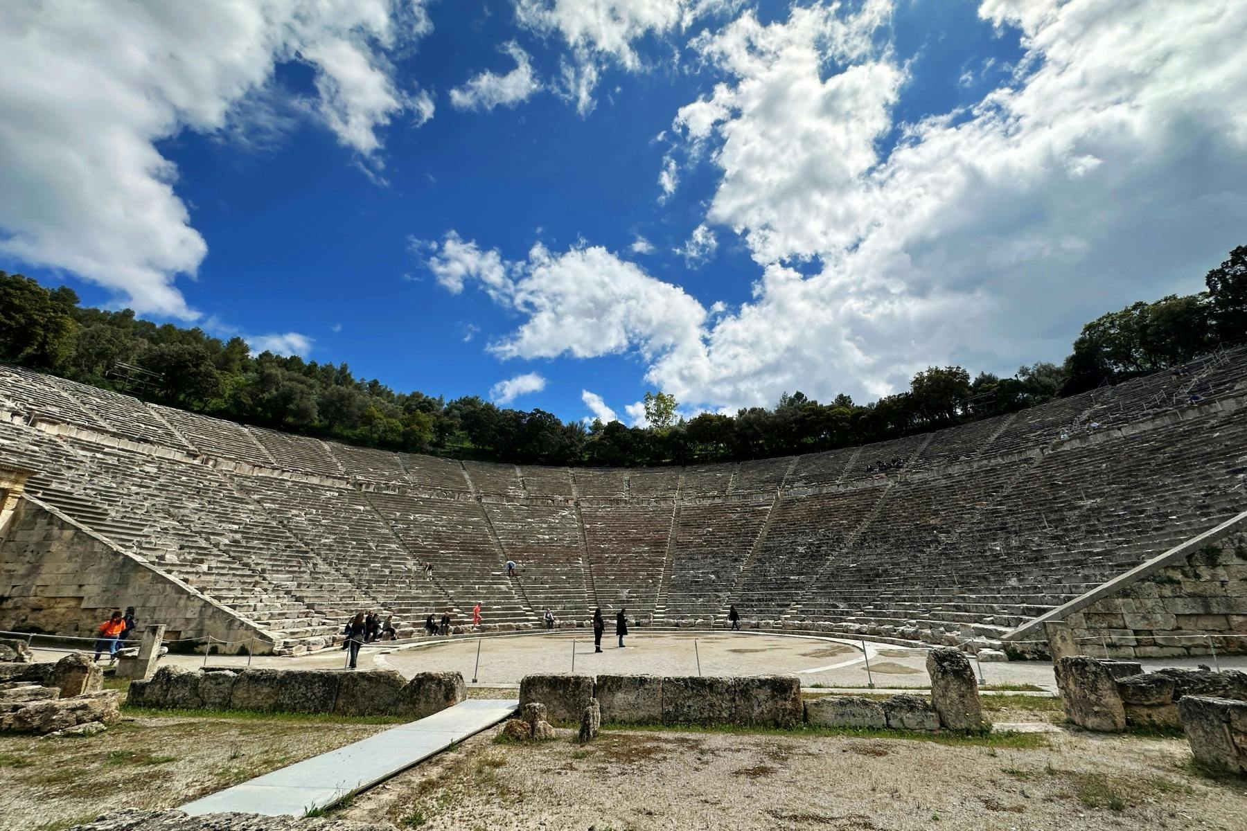 A panoramic view of the ancient Epidaurus theater with stone seats and a round orchestra under a blue sky.