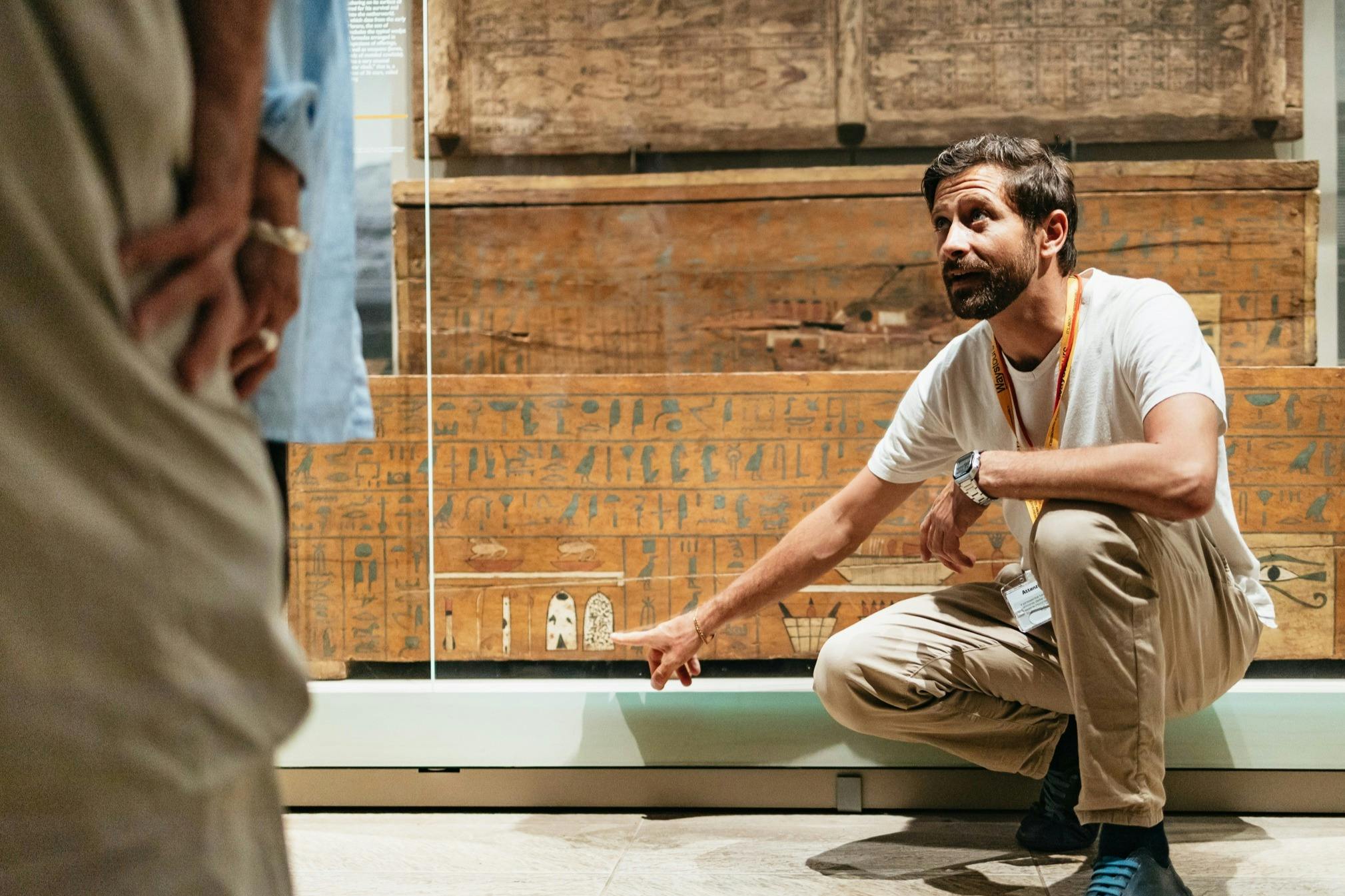A man crouches and gestures toward ancient carvings or inscriptions on a wooden artifact in a museum setting.