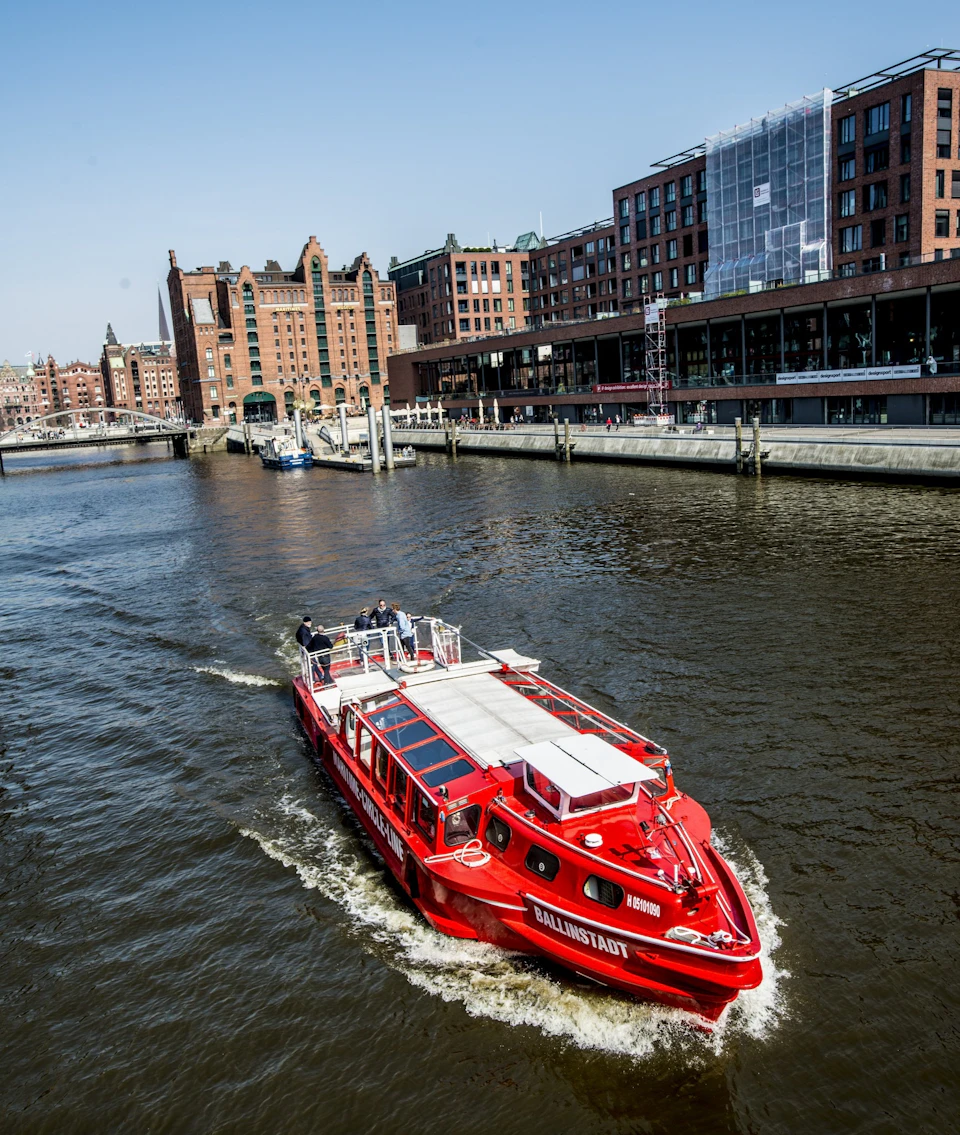 A red boat sails down a river flanked by historic brick buildings and modern structures under a clear blue sky.