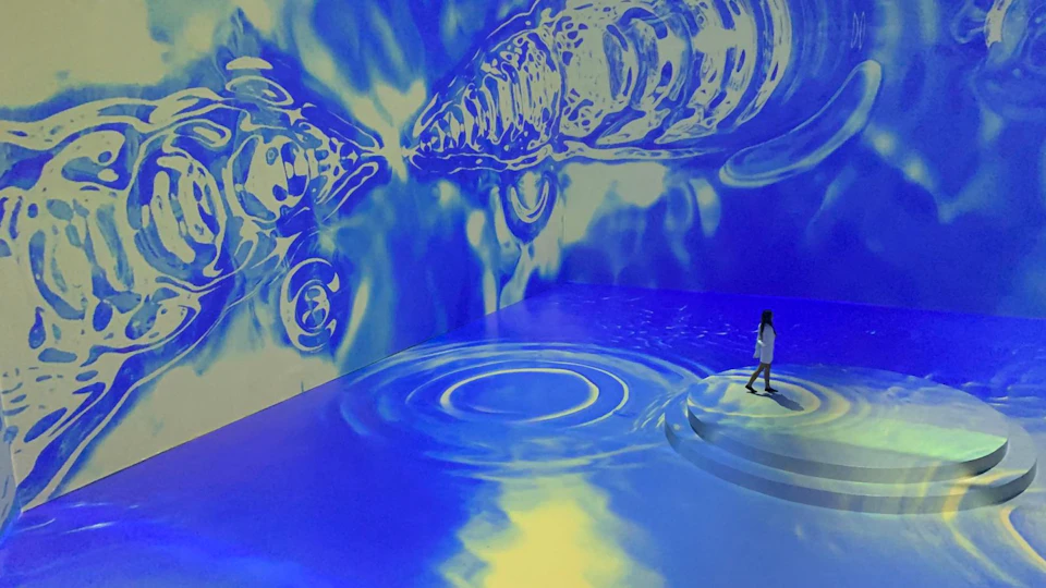 A person stands in a room with immersive blue and yellow lighting and projected patterns resembling water ripples and waves.