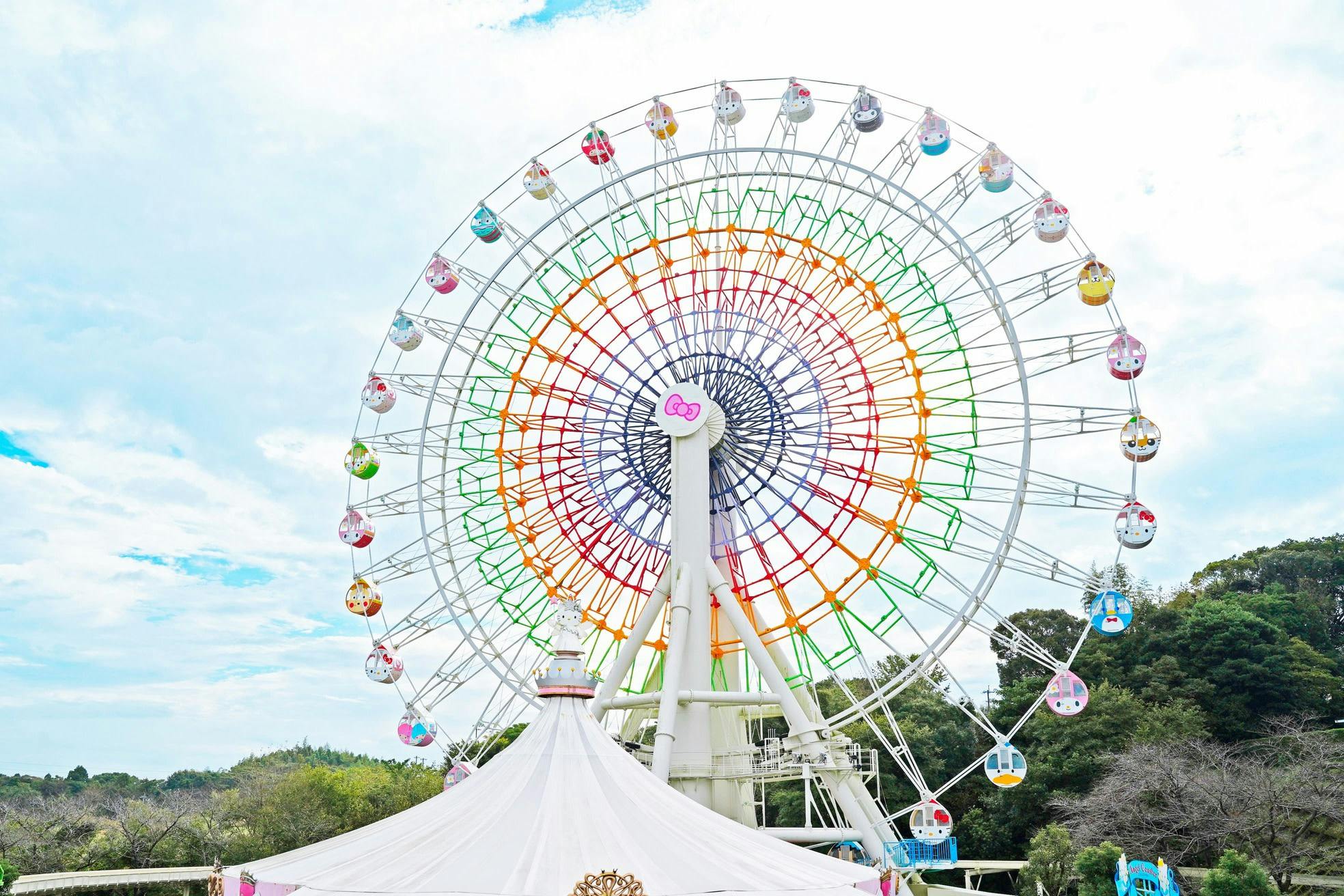 A colorful ferris wheel with themed gondolas against a partly cloudy sky, with trees and a white tent in the foreground.