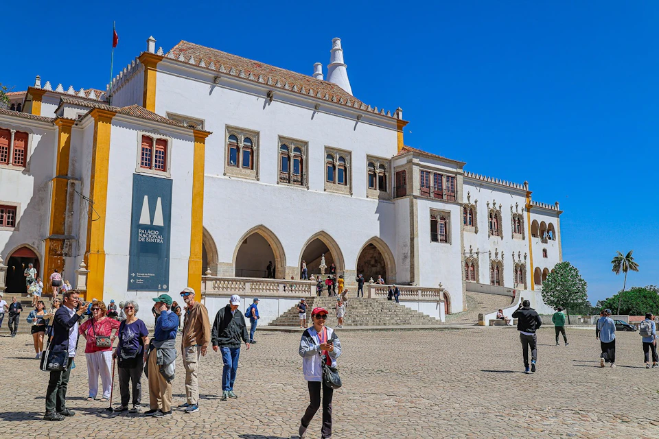 A historic white building with arched windows and chimneys, with visitors walking on a cobblestone courtyard under a clear sky.