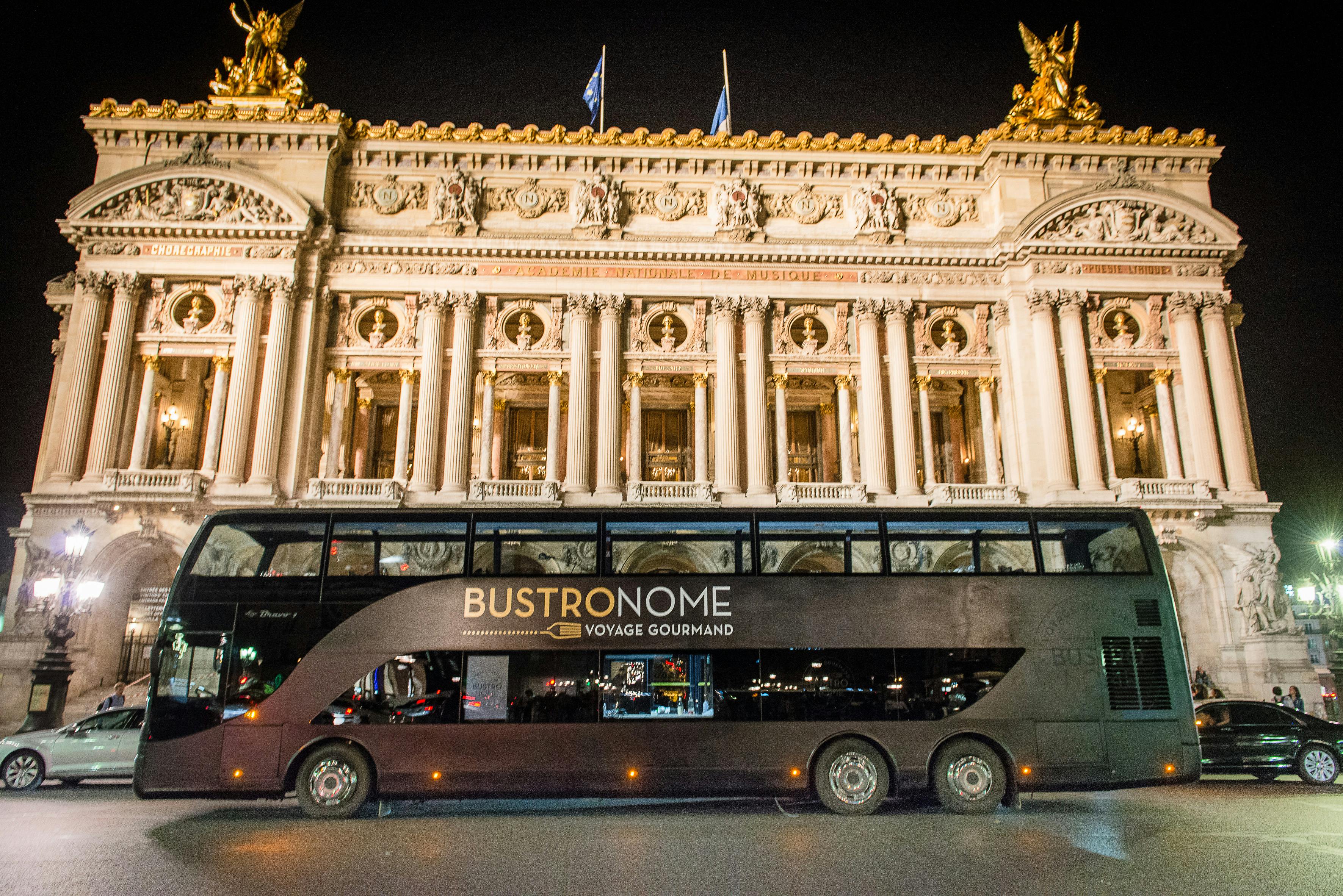 夜间，一辆名为「Bustronome Voyage Gourmand」的双层巴士停在装饰华丽的建筑前。