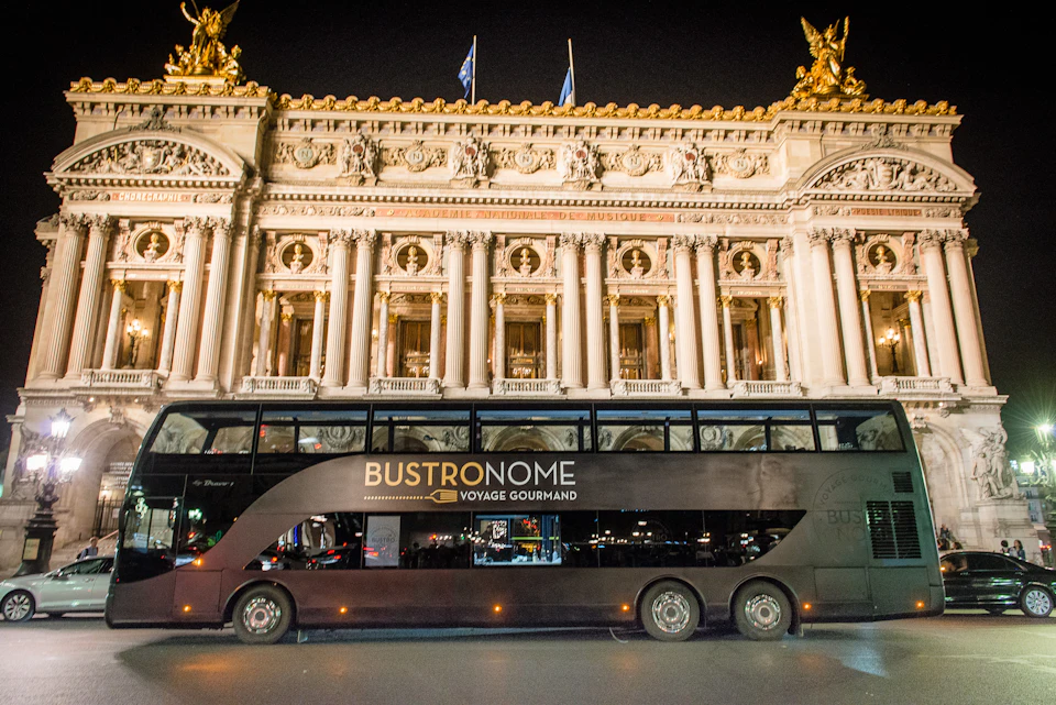 A double-decker bus labelled "Bustronome Voyage Gourmand" is parked in front of an ornately decorated building at night.