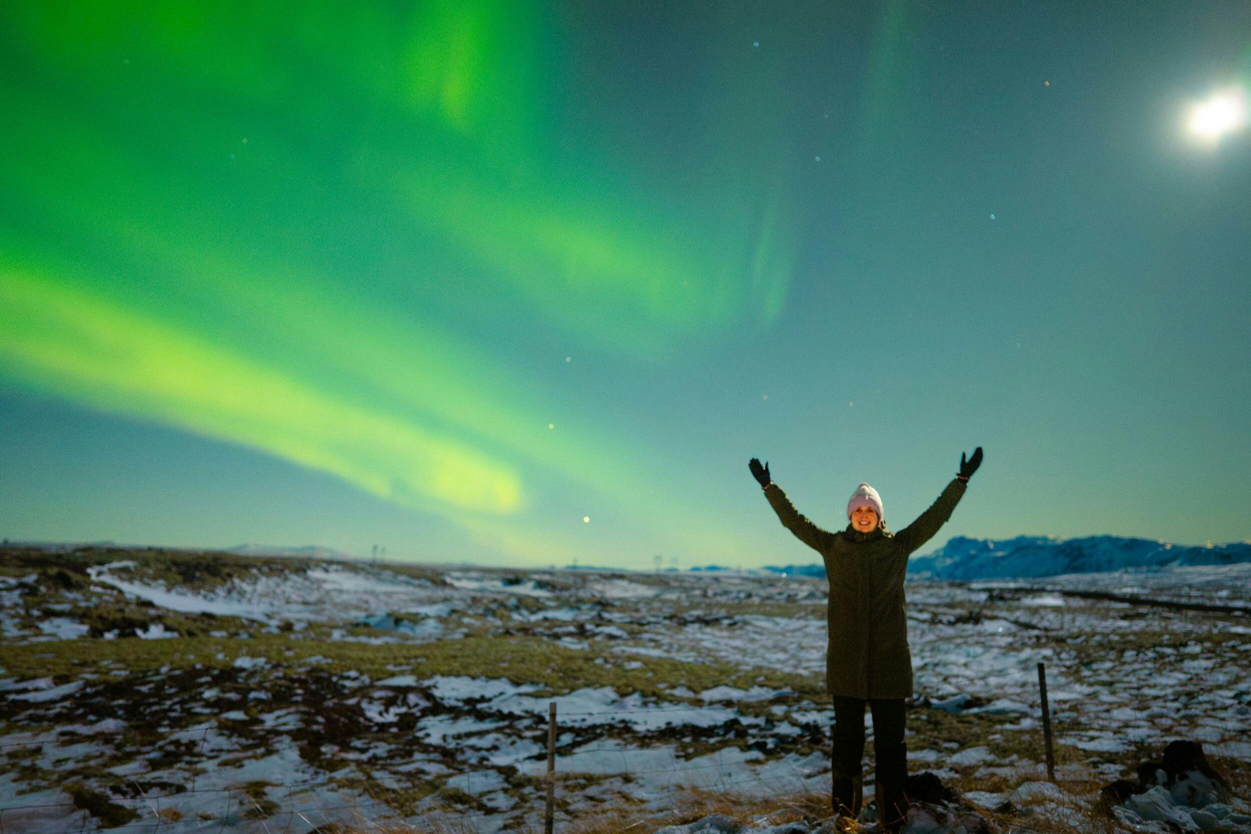 A person in winter clothing stands with arms raised under a vivid green aurora in a snowy landscape at night.