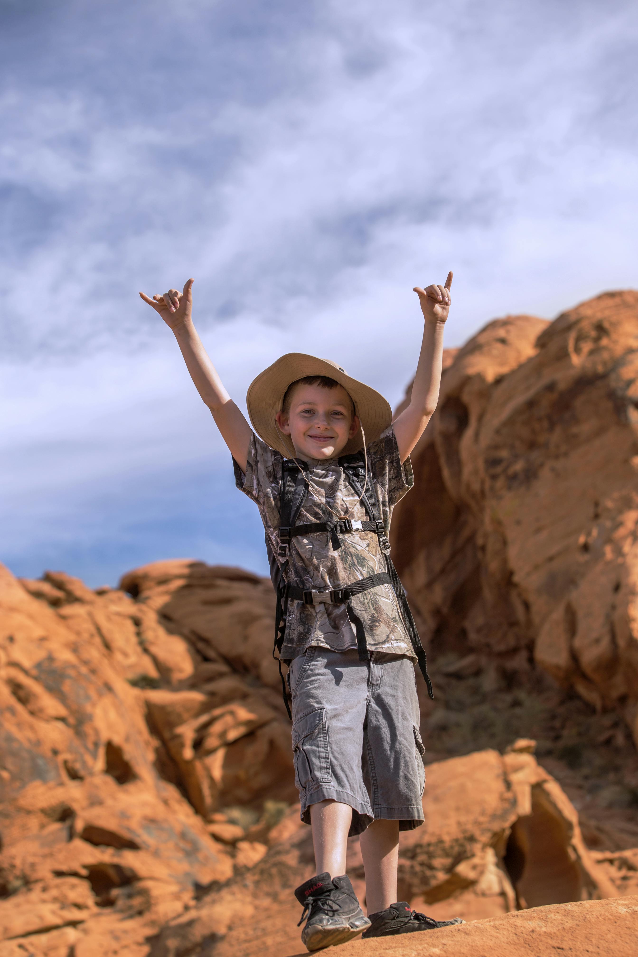 Smiling boy in a camo shirt and wide-brimmed hat stands in front of red rock formations, arms raised, under a blue sky with clouds.
