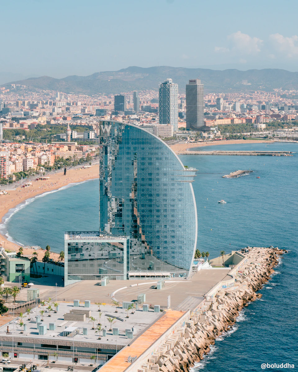 Aerial view of a coastal city with a prominent sail-shaped hotel, a long beach, and urban buildings in the background.
