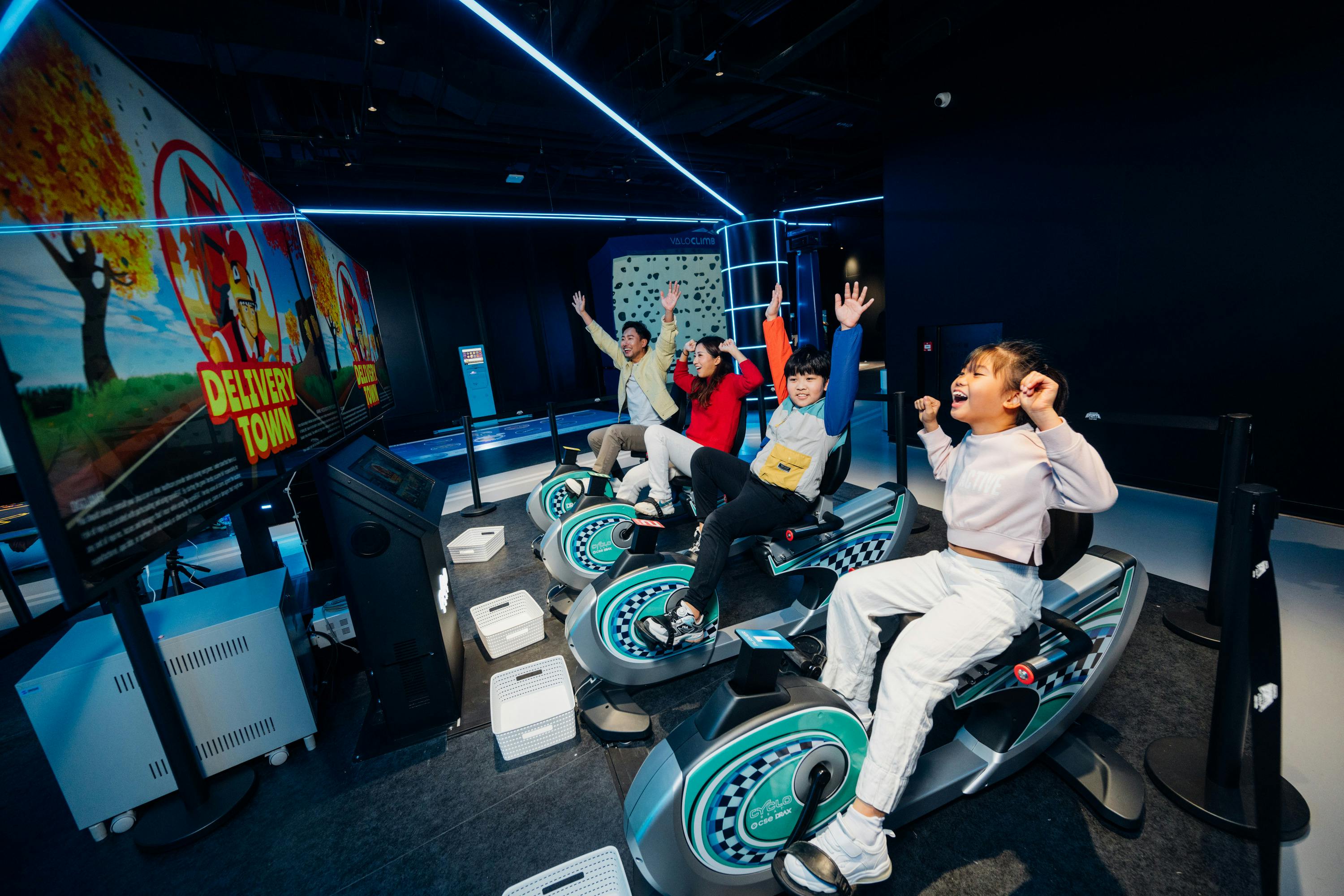 Children enthusiastically riding stationary bikes in a futuristic, neon-lit indoor setting.