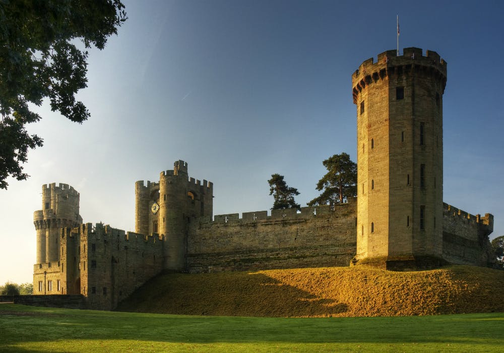 Stone castle with tall, round towers against a clear sky, surrounded by a grassy courtyard and trees in the background.