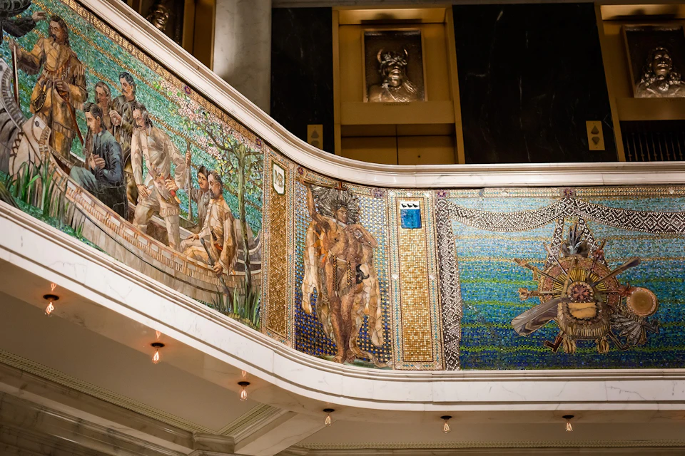 Intricately detailed mosaic mural on a curved wall in a building, depicting various human figures and symbolic scenes.