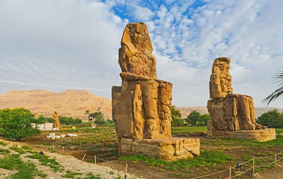 Two large ancient stone statues sit in a grassy landscape with mountains and a partially cloudy sky in the background.