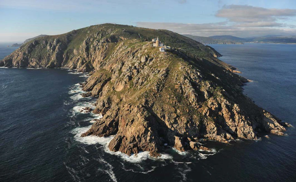 Rocky coastline with crashing waves and a lighthouse on a hilly promontory extending into the ocean.