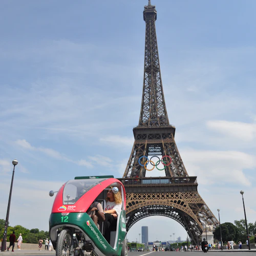 A three-wheeled pedicab with passengers in front of the Eiffel Tower, which has Olympic rings displayed in its structure.