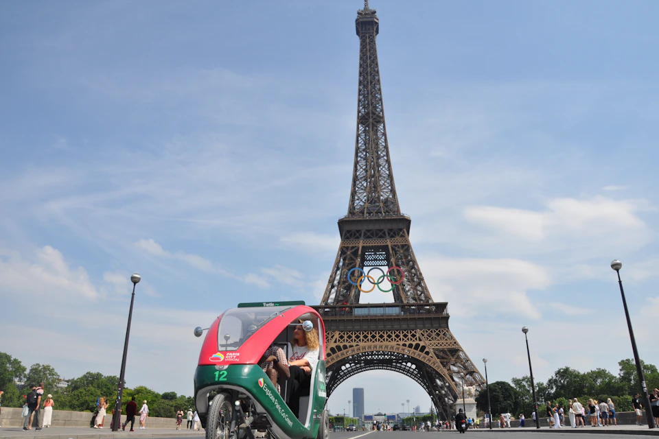 A three-wheeled pedicab with passengers in front of the Eiffel Tower, which has Olympic rings displayed in its structure.
