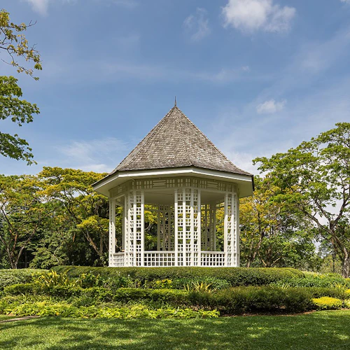 Ein weißer Pavillon mit Schindeldach ist umgeben von gepflegtem Gras und üppigen Bäumen unter einem strahlend blauen Himmel.