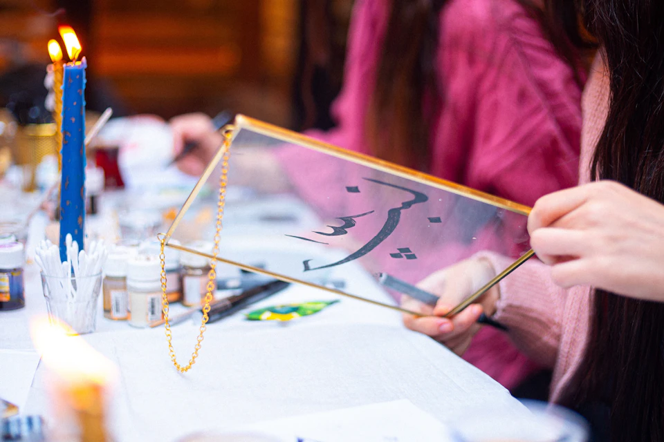 A person holding a rectangular glass frame with decorative calligraphy, seated at a table with art supplies.