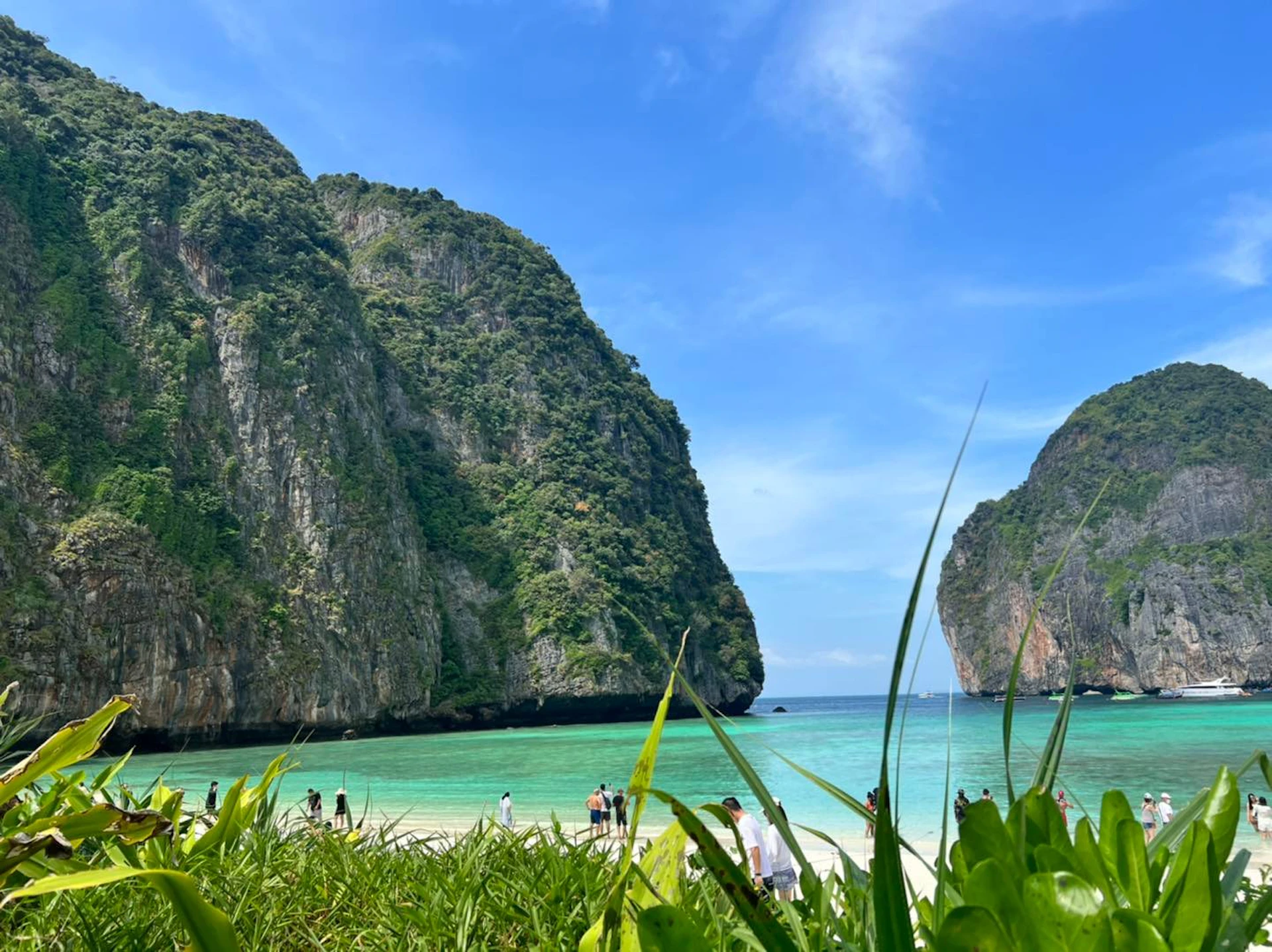 Beach scene with people in the distance, turquoise water, and tall, green cliffs under a blue sky with light clouds.
