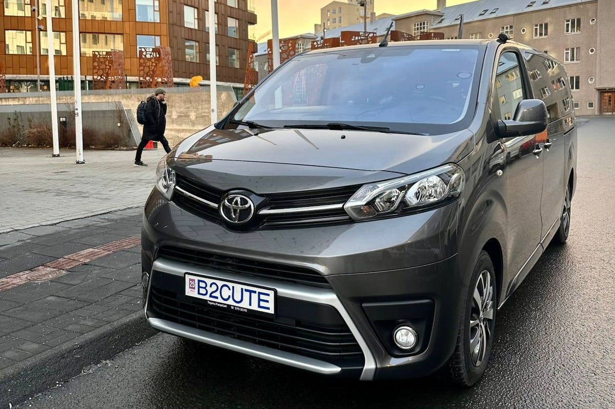 市街地に専用ナンバープレート「B 2 CUTE」を駐車したグレーのトヨタミニバン。 歩行者がバックグラウンドで表示されます。