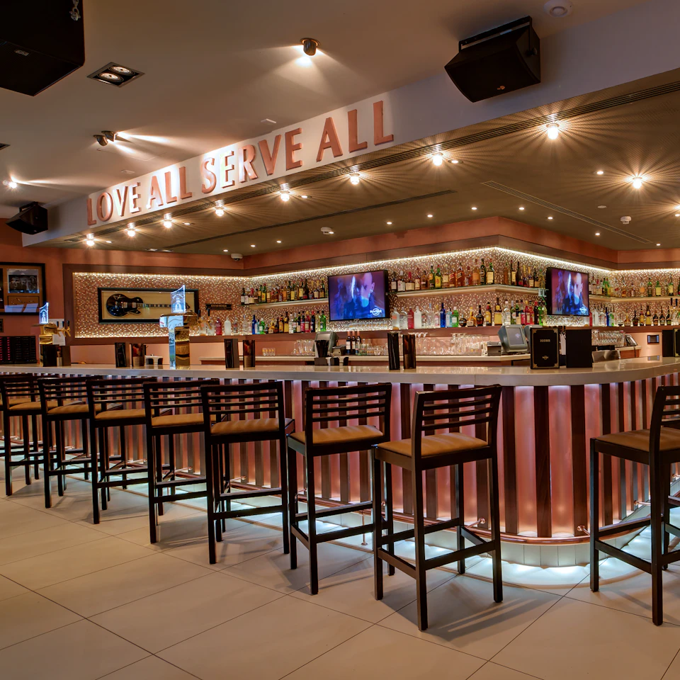A bar with a long counter, high chairs, illuminated shelves of liquor bottles, and two TVs. Sign above reads "LOVE ALL, SERVE ALL."