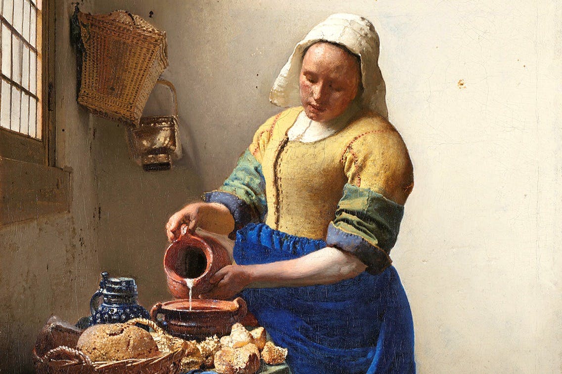 A woman in a white headscarf pours milk from a jug into a bowl on a table with bread.