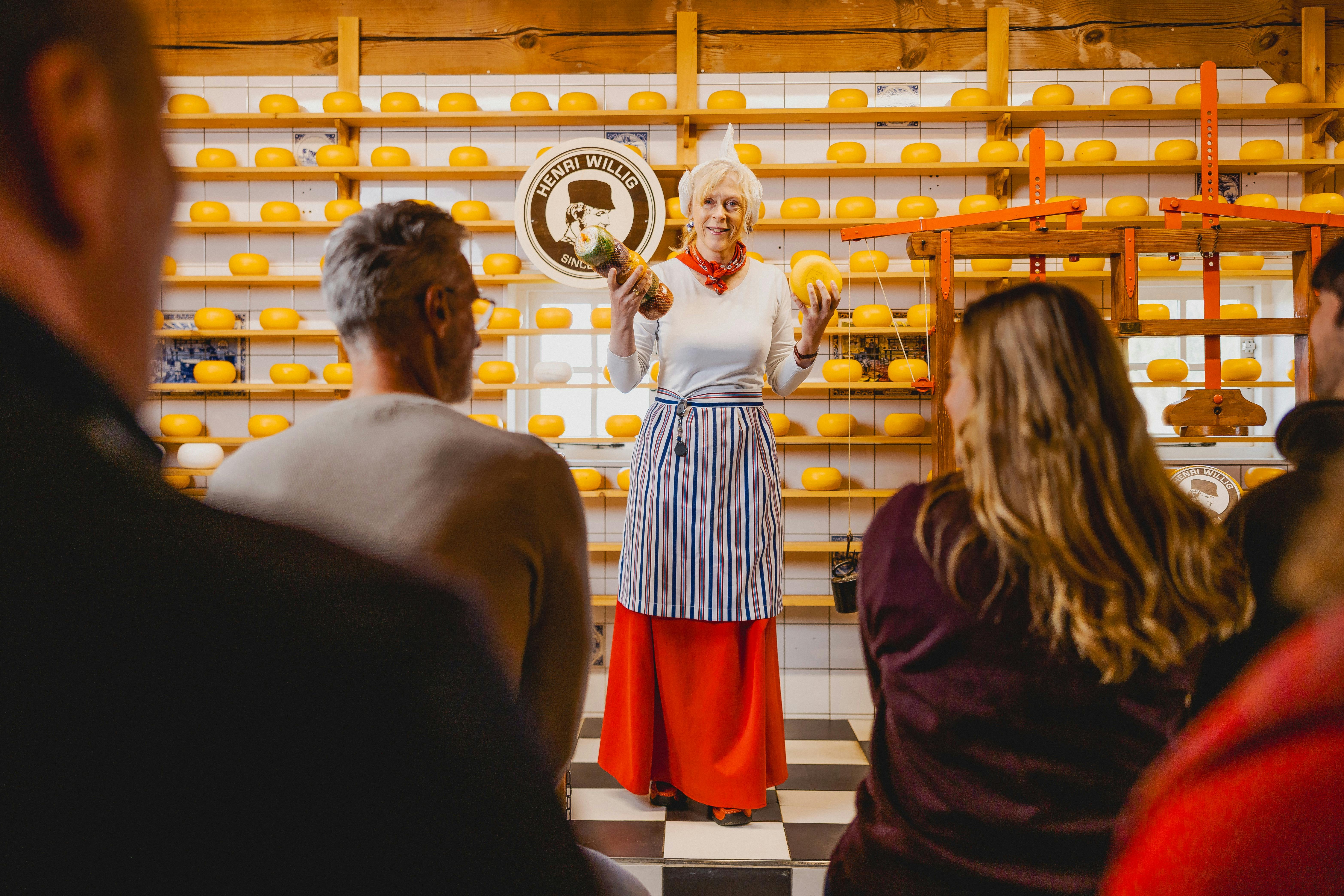 Μια γυναίκα με ποδιά στέκεται μπροστά από ράφια γεμάτα με τροχούς τυριού, κρατώντας και δείχνοντας τυριά σε ένα ακροατήριο.