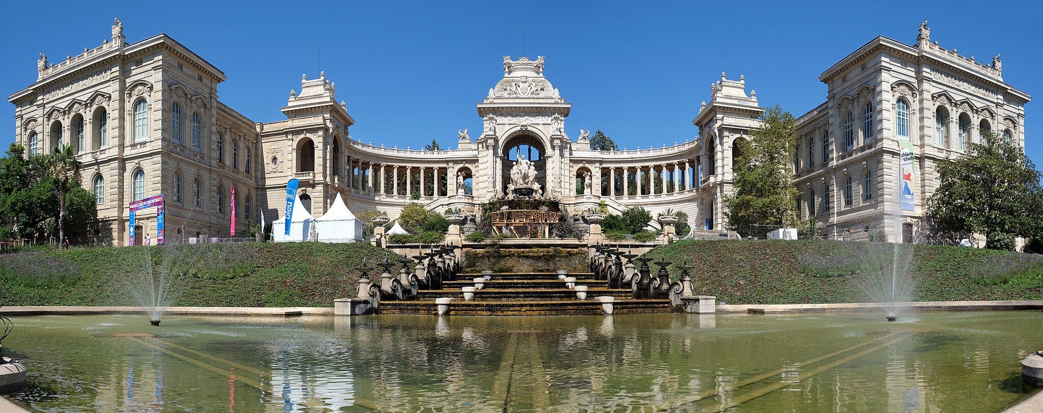 Ornate building with two symmetrical wings, a central arch, fountains, statues, and a reflecting pool under a clear blue sky.