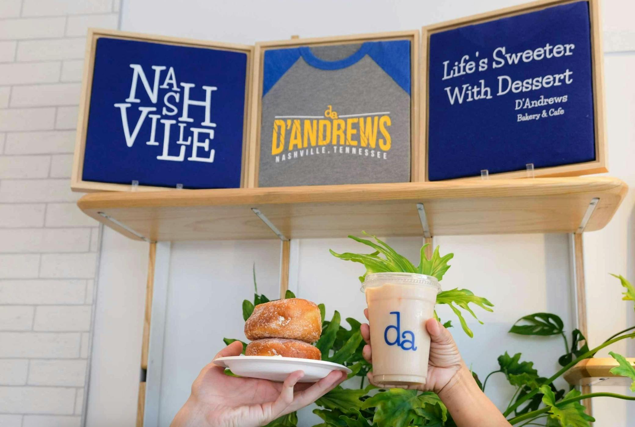 Mani che tengono in mano un piatto di ciambelle e una bibita ghiacciata davanti alla merce di D'Andrews Bakery & Cafe con insegne.
