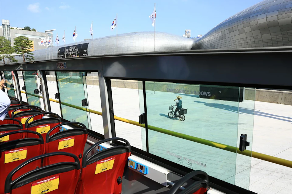 Interior of a bus with red seats, a cyclist outside on a plaza, modern architecture, and flags flying in the distance.