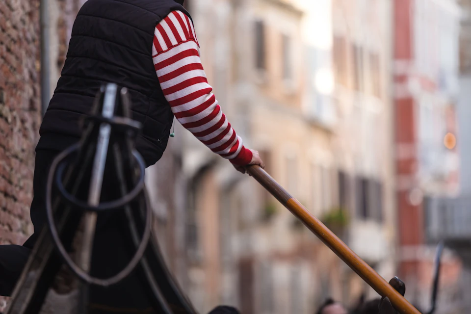 A person in a striped shirt and vest rowing a gondola with a wooden oar in a city with blurred buildings in the background.