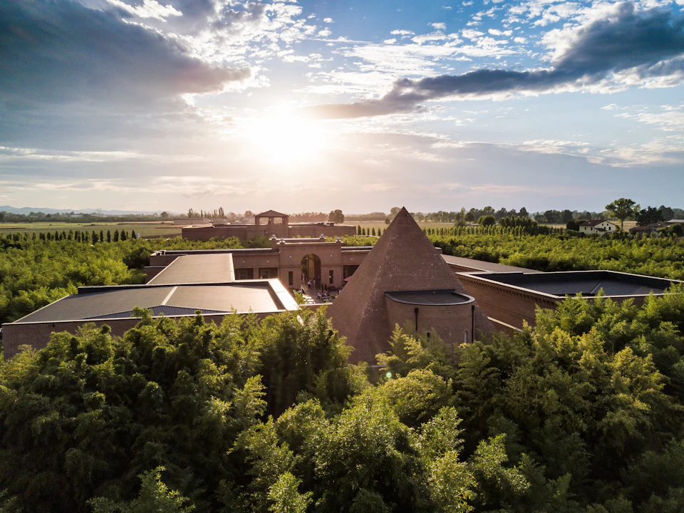 A brick pyramid structure surrounded by greenery and other buildings under a partly cloudy sky with the sun setting in the distance.
