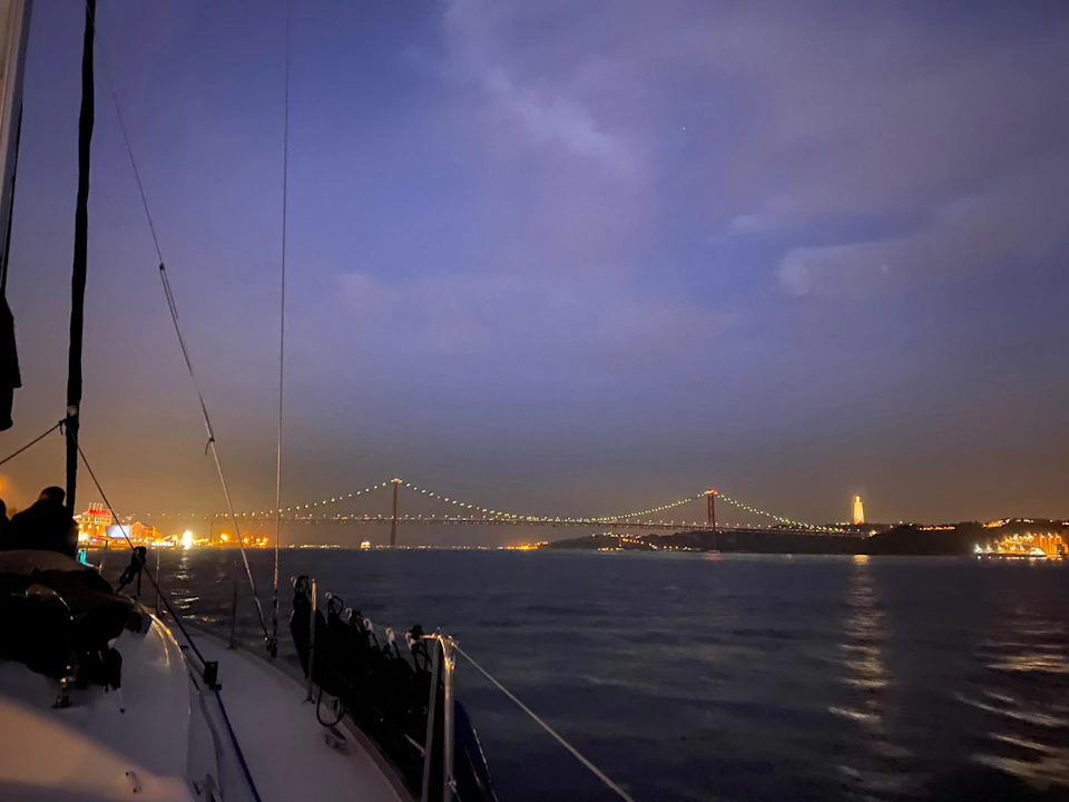 View from a boat at night, looking towards a lit suspension bridge over a body of water with city lights in the background.