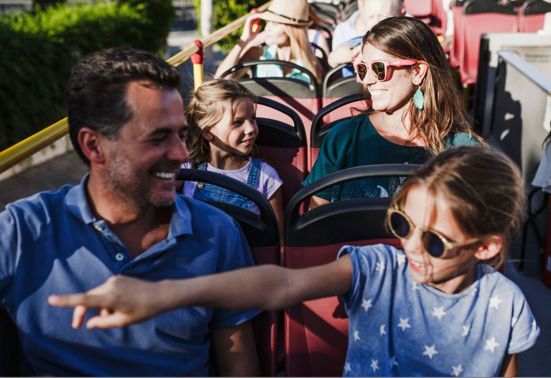 Un gruppo di persone sorride e parla sul ponte superiore di un autobus scoperto; un bambino con gli occhiali da sole indica eccitato.