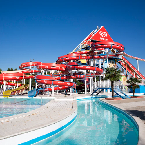 Vattenpark med flera röda, gula och blå vattenrutschbanor, pooler och palmer, under en klarblå himmel.