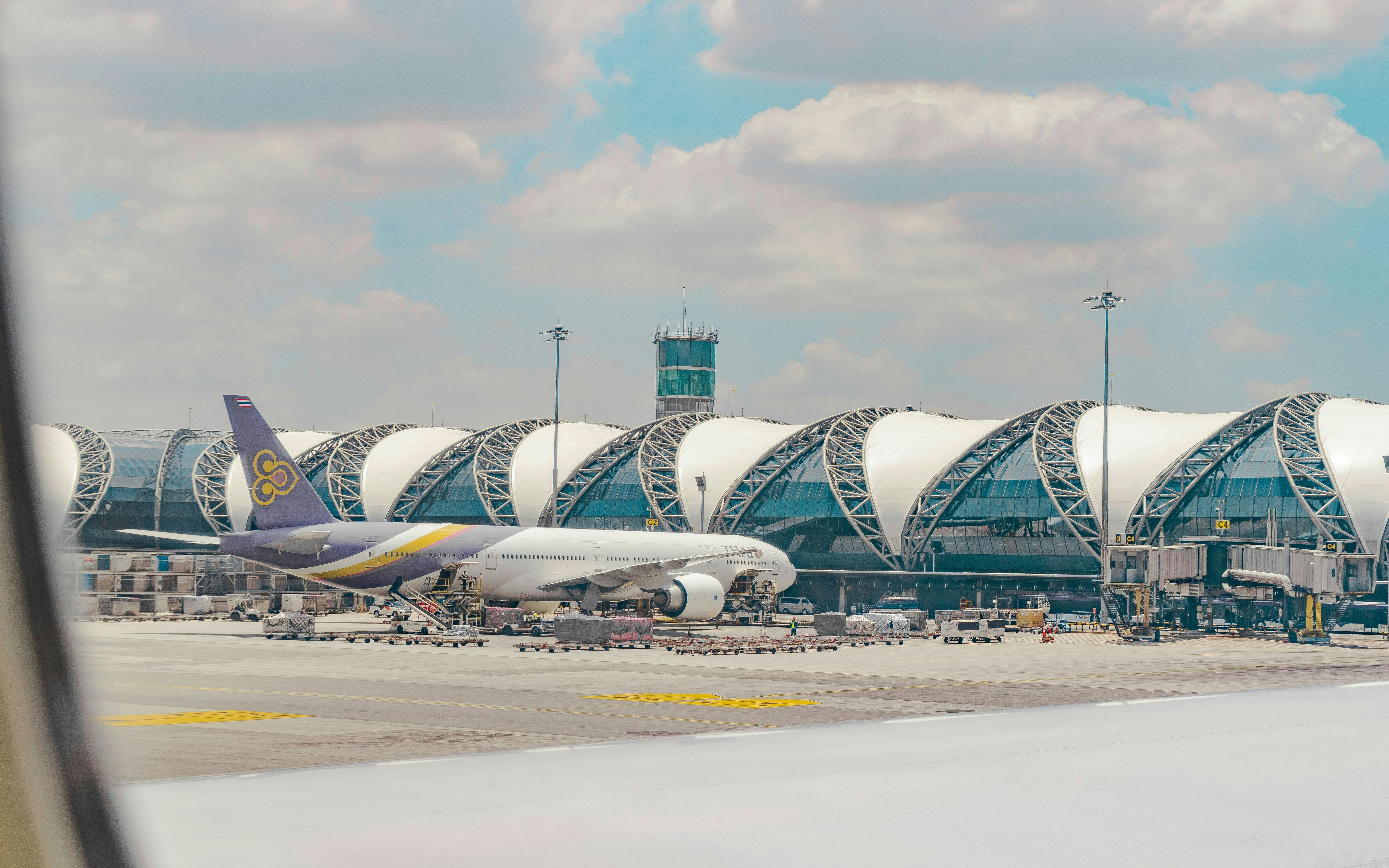 Un gros avion est stationné dans un terminal d'aéroport moderne avec un toit arqué distinctif et une tour de contrôle sous un ciel partiellement nuageux.