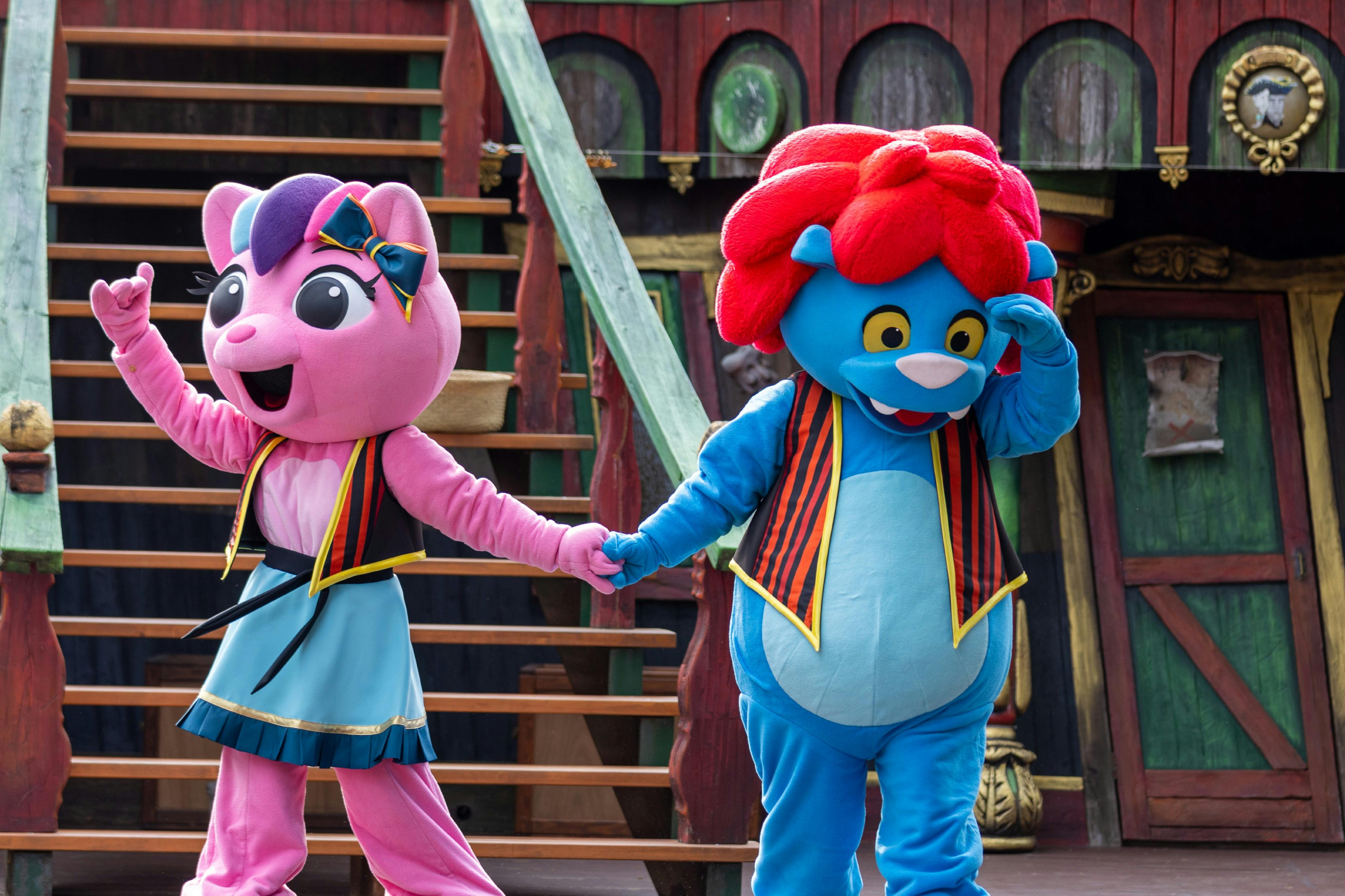Two colorful mascots, a pink cat and a blue bear, wearing costumes, hold hands on a set resembling a pirate ship.