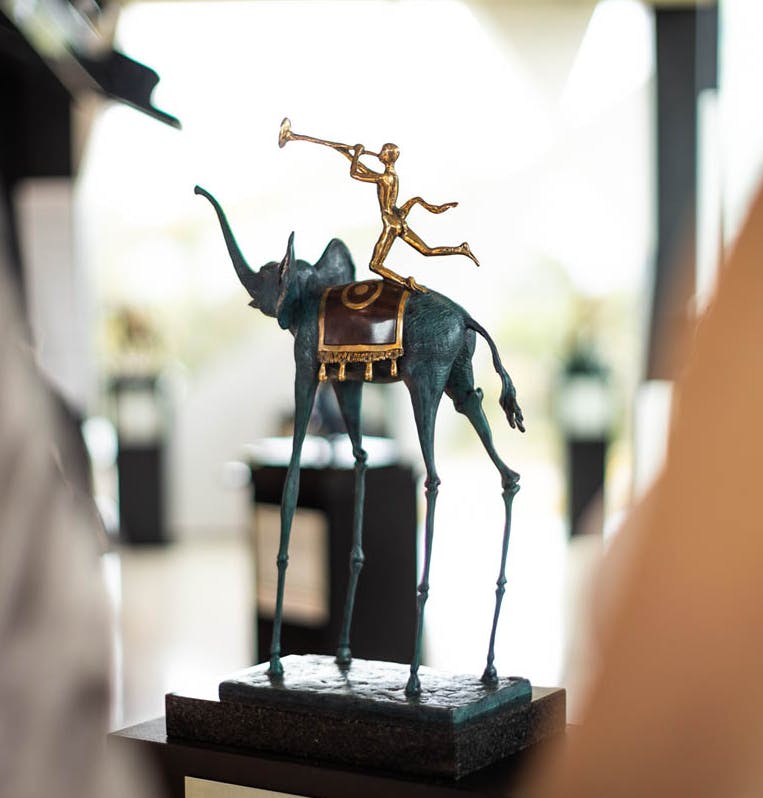 Χάλκινο γλυπτό ενός λεπτού, με μακριά πόδια ελέφαντα με μια σέλα, που μεταφέρει μια φιγούρα που παίζει τρομπέτα.
