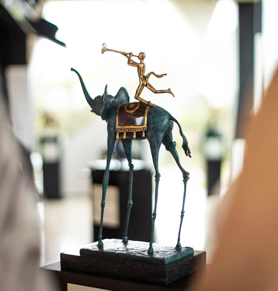 Bronze sculpture of a thin, long-legged elephant with a saddle, carrying a figure playing a trumpet.